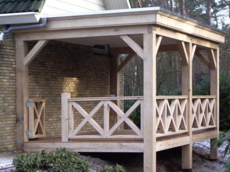 Houten veranda met balustrade en ondersteunende balken, naast een bakstenen muur.