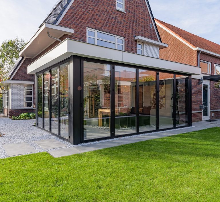 Moderne glazen uitbouw aan een woning met een groene tuin.
